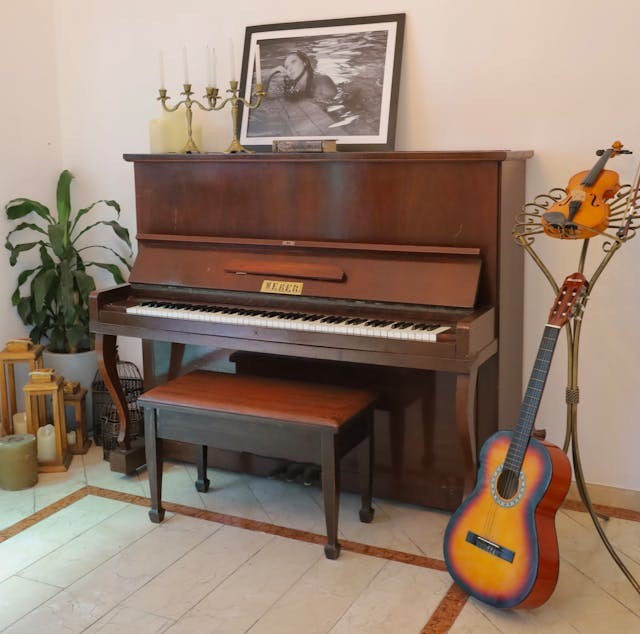 Hnědé pianino s klavírní stoličkou stojící u vnitřní zdi bytu. Vedle stojí klasická kytara.